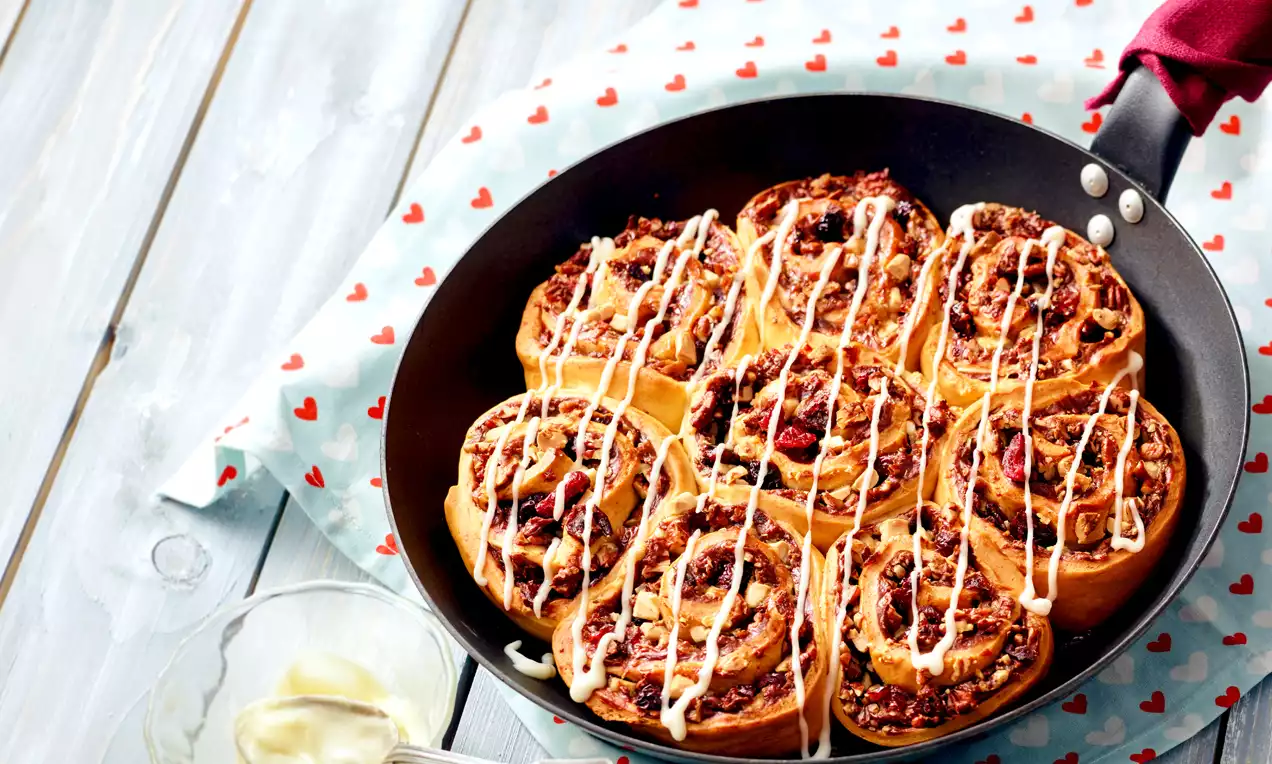 Goldbraune Zimtschnecken mit karamellisierter Nussfüllung und weißem Zuckerguss in einer schwarzen Pfanne auf einem hellblauen Stoff mit Herzen