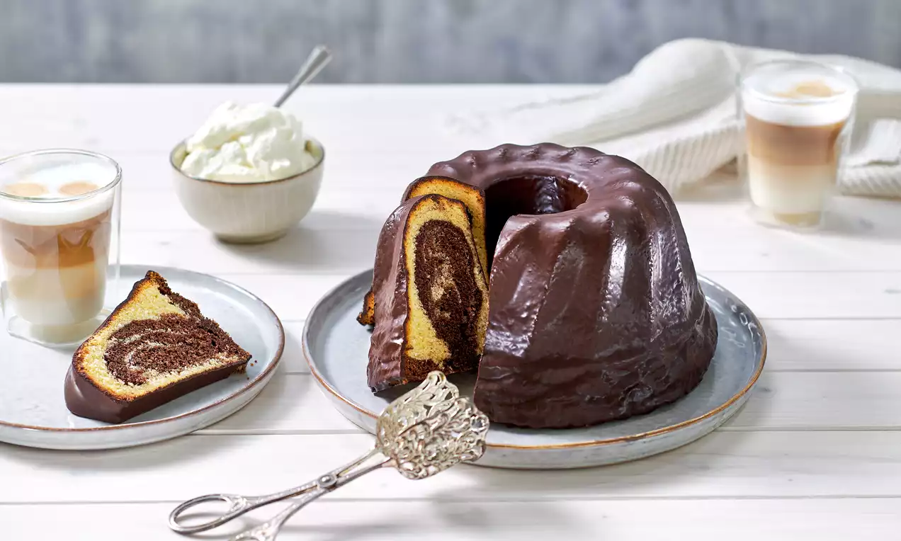 Runder Marmorkuchen mit glänzender Schokoladenglasur auf einer Platte, angeschnitten mit sichtbarer Marmorierung, umgeben von Sahne und Kaffee