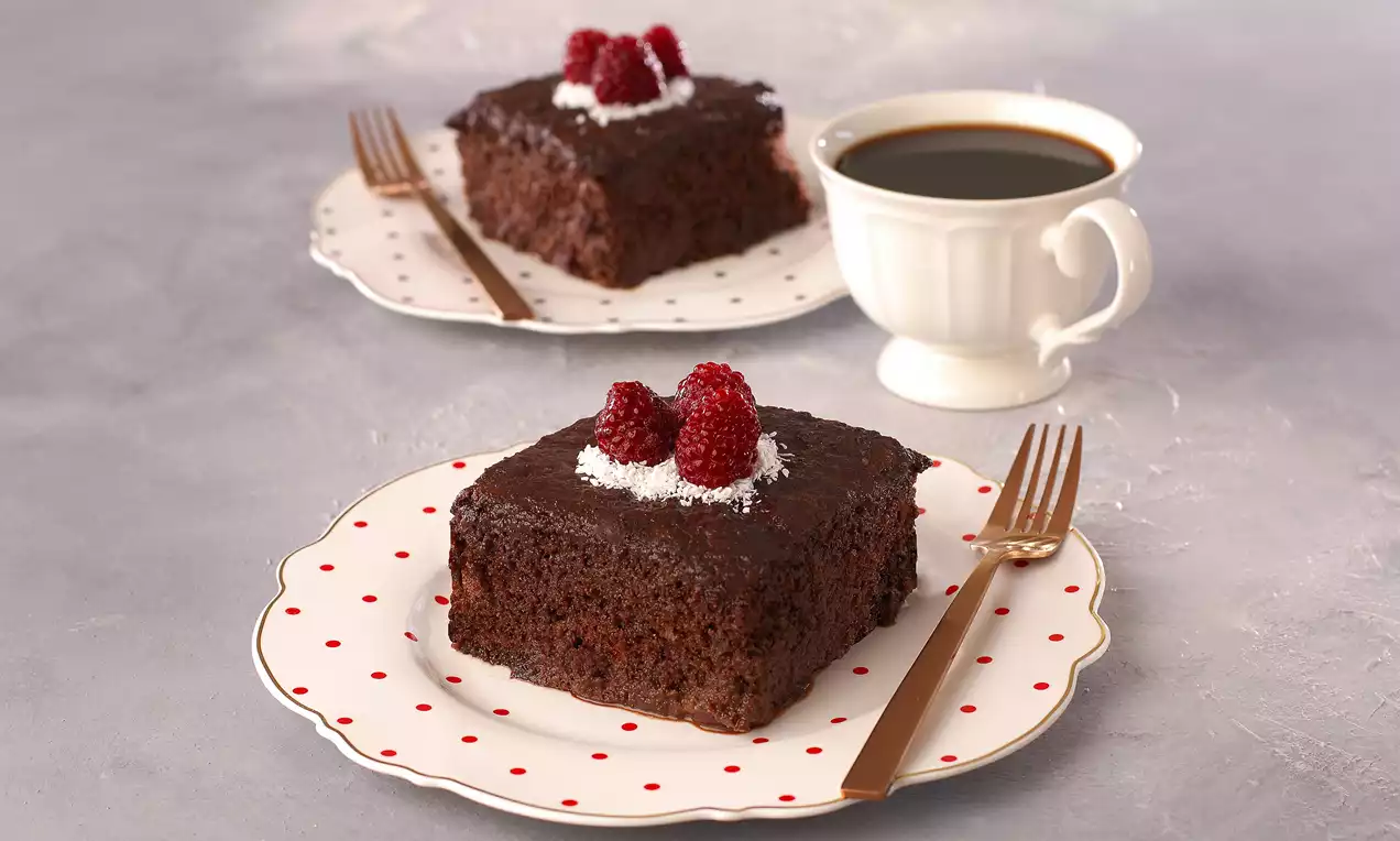 Yoğun çikolatalı ıslak kek, üzerine hindistan cevizi serpiştirilmiş ve frambuazlarla süslenmiş desenli tabakta bronz çatal ile sunulmuş