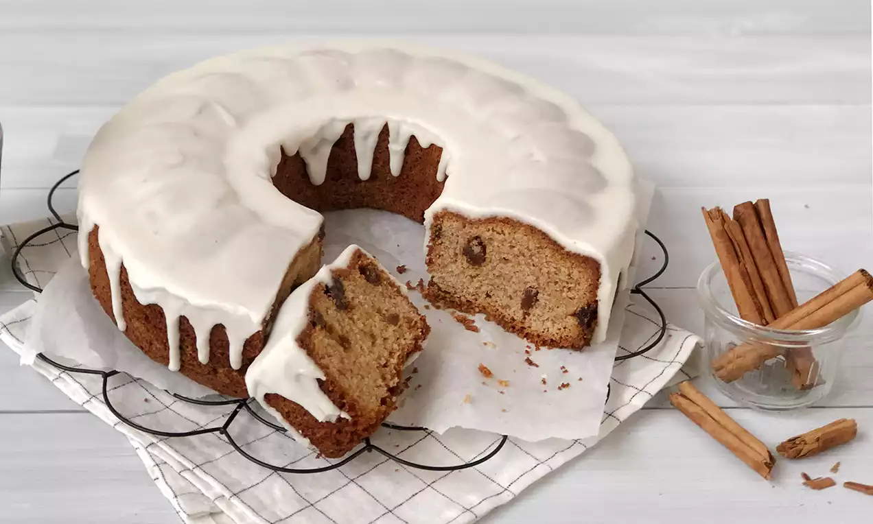 Zimtkuchen in Kranzform mit glatter weißer Glasur, innen mit braunen Rosinen, auf Backpapier mit Zimtstangen dekoriert