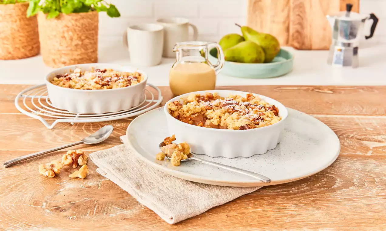 Birnen-Crumble mit goldbraunen Streuseln in einer weißen, wellenförmigen Auflaufform, angerichtet auf einem weißen Teller mit Stoffserviette