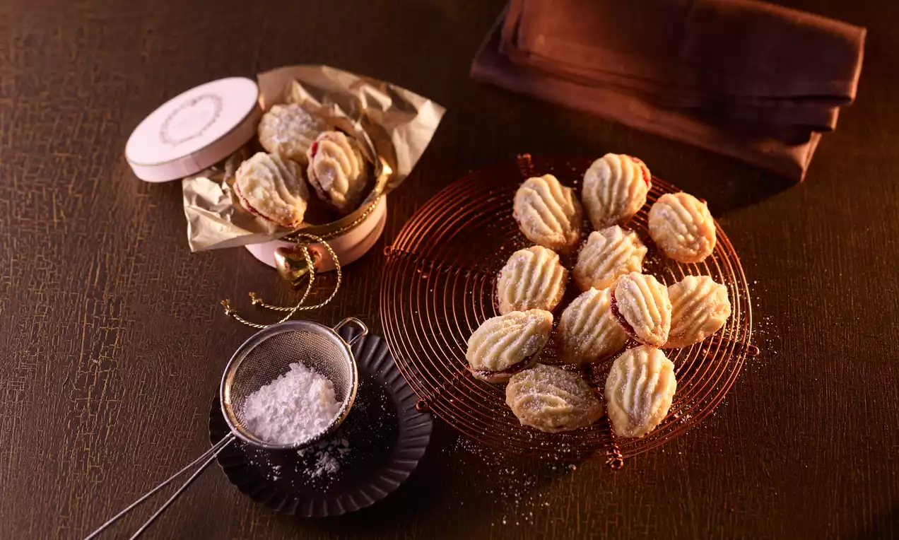 Biscotti dorati con forma ondulata e crema al centro, disposti su una griglia rossa accanto a una scatola decorata e un setaccio con zucchero