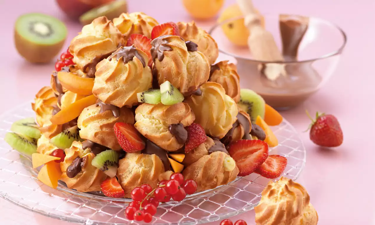 Goldbraune Profiteroles mit glänzender Schokoladenglasur, umgeben von frischen Sommerfrüchten auf einer gläsernen Servierplatte