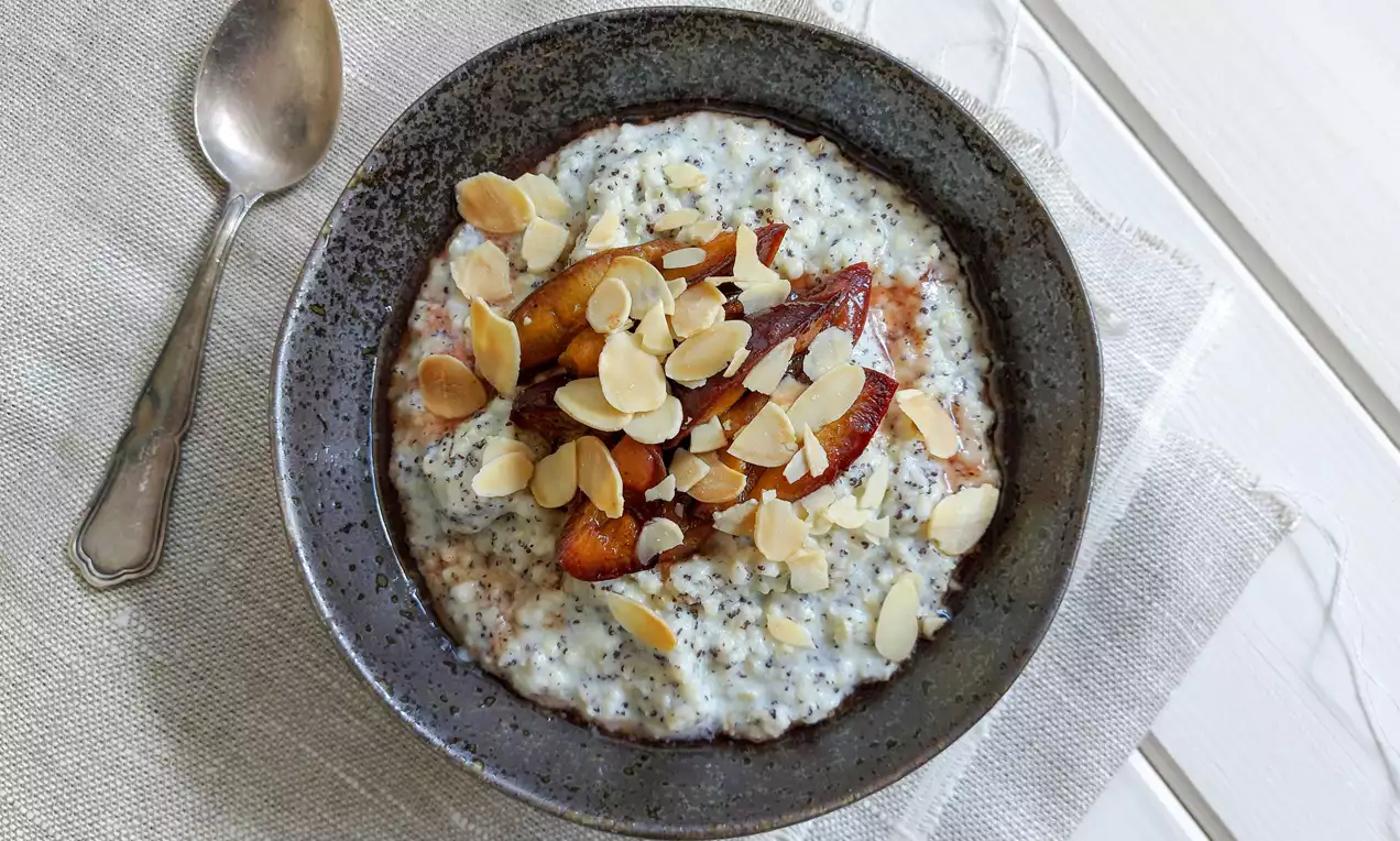 Maková jáhlová kaše světle šedé barvy s jemnou texturou, zdobená pečenými švestkami do hněda a plátky mandlí v keramické misce