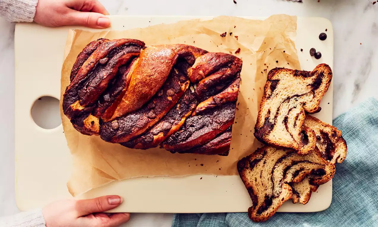 Golden-brown babka loaf with dark chocolate swirls, a shiny twisted crust, and sliced pieces set on parchment paper on a white cutting board
