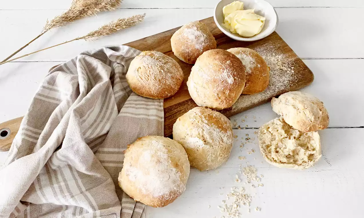 Pyöreät sämpylät vaaleanruskeita ja kevyesti jauhotettuja, aseteltuina puiselle leikkuulaudalle, vierellä voita kulhossa ja pellavakangasta