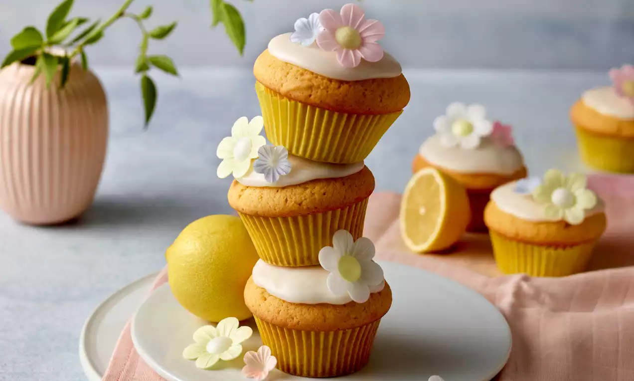 Zitronen-Muffins in gelben Förmchen mit weißem Zuckerguss und zarten, bunten Blüten dekoriert auf einem weißen Teller mit frischer Zitrone