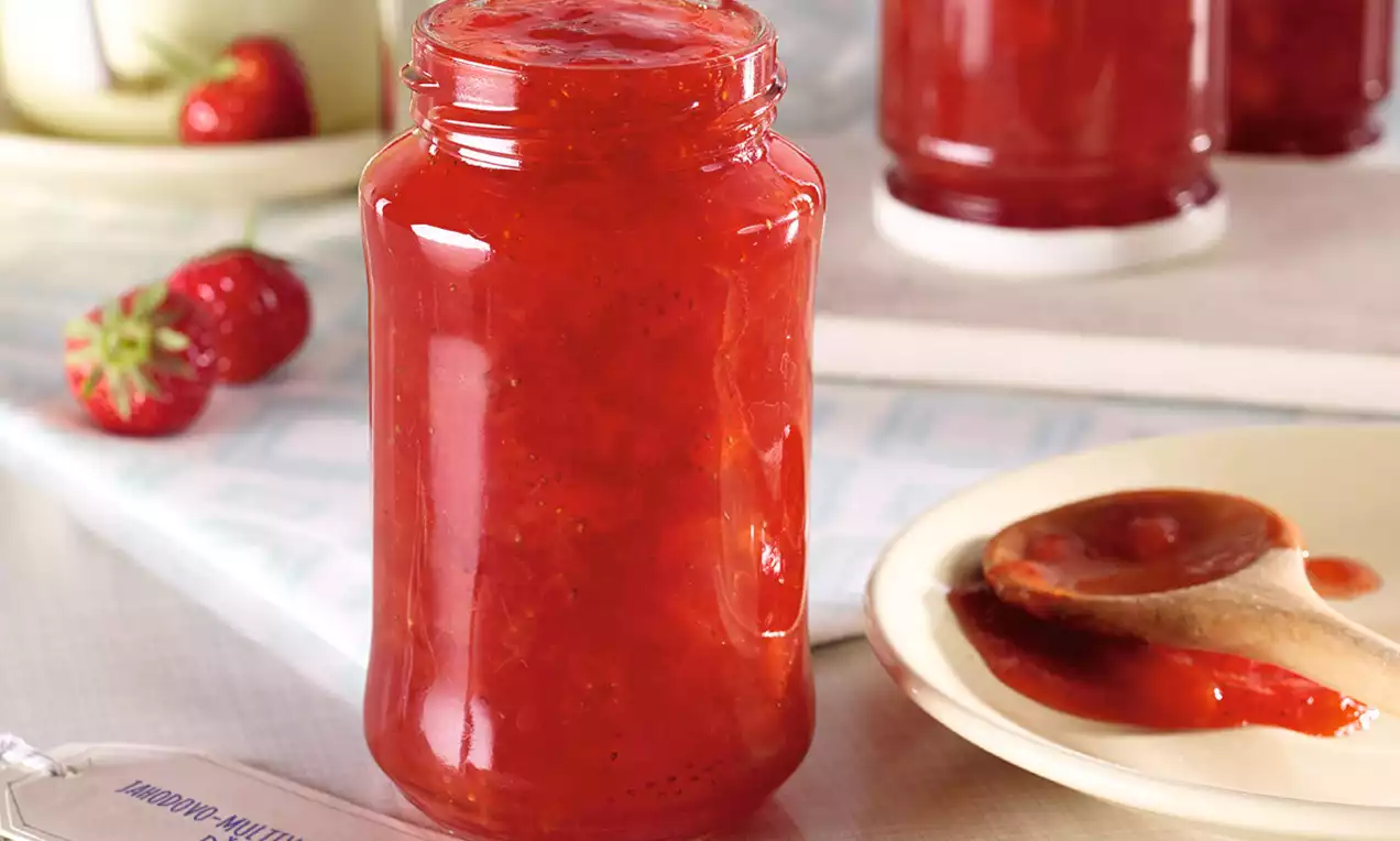 Sklenice s jasně červeným džemem se zrníčky, lesklý povrch, vedle dřevěná lžička s džemem na béžovém talířku, v pozadí čerstvé jahody