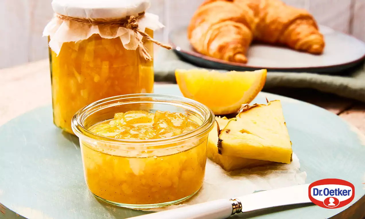 Sklenice ananasovo-pomerančové marmelády s viditelnými kousky ovoce vedle řezů ananasu a pomeranče, croissantu a nože na světle modrém podnosu