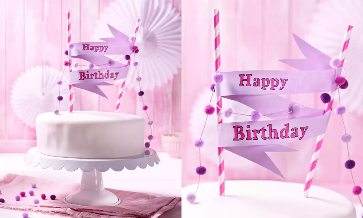 Kleine runde Torte mit weißem Fondant auf Tortenplatte, dekoriert mit lila Papierwimpeln und bunten Pompoms in Rosa- und Lilatönen