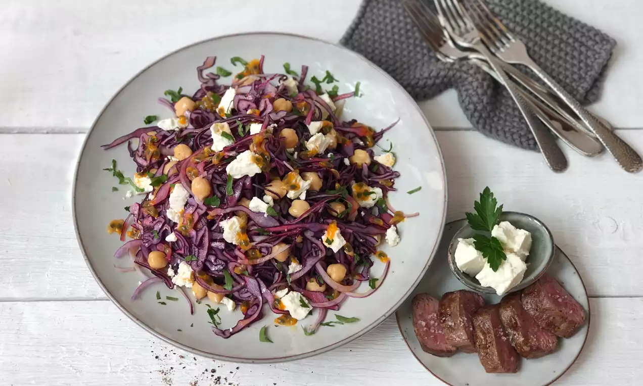 Rotkohl-Kichererbsen-Salat mit weißem Feta und frischen Kräutern auf hellem Teller, daneben gegrilltes Fleisch und Feta-Schälchen