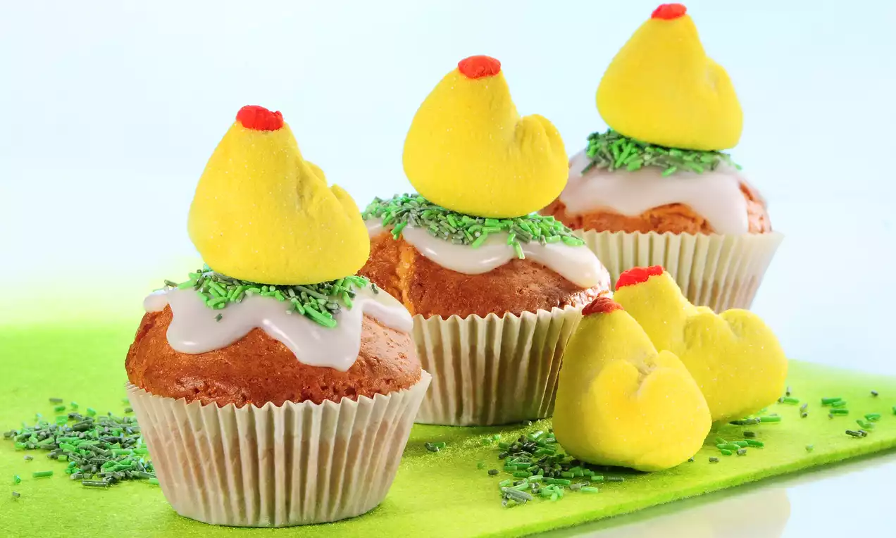 Babeczki w białych papilotkach z lukrem i zielonymi wiórkami, zwieńczone żółtymi piankowymi kurczakami z czerwonymi akcentami na pastelowej serwetce