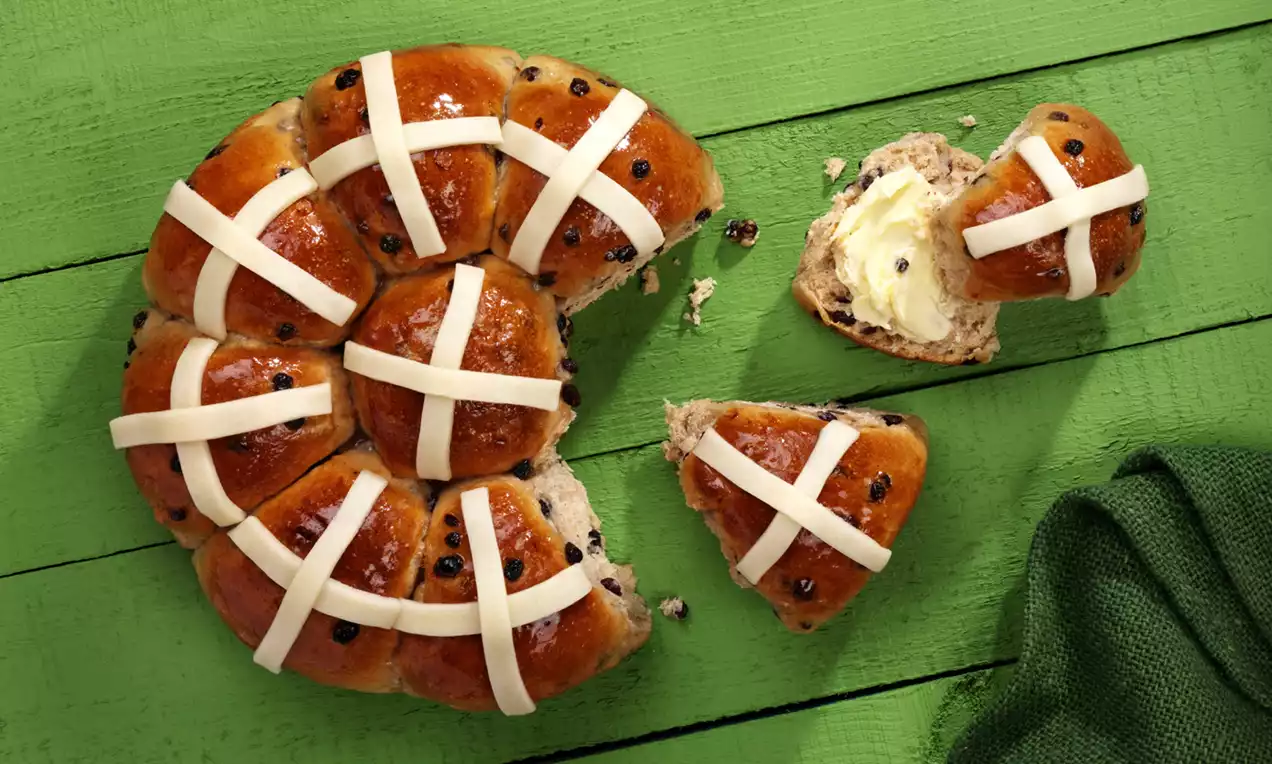Golden-brown tear-and-share hot cross buns with white icing crosses arranged in a ring on a green wooden table with a cloth nearby