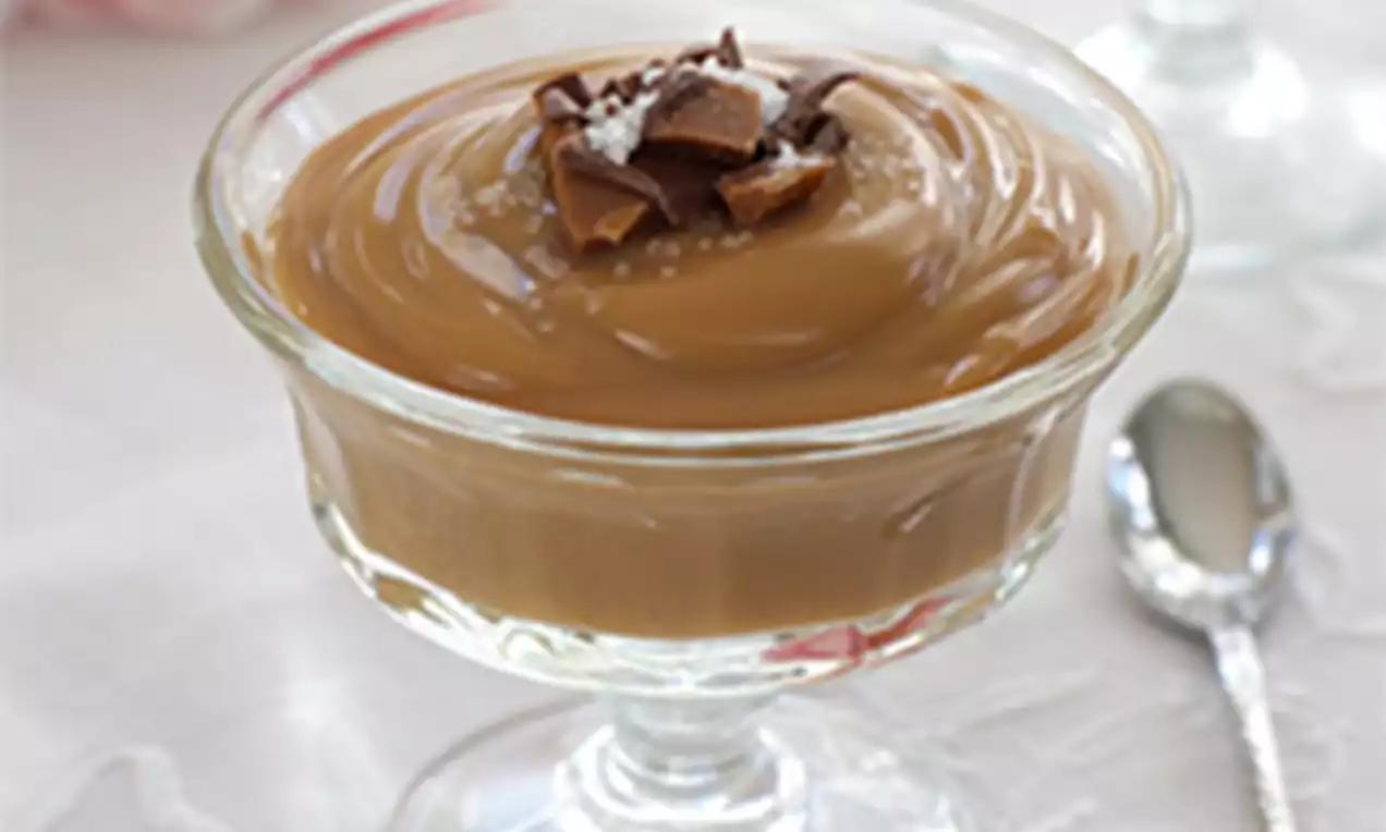 Glass dessert dish filled with glossy caramel-colored sticky toffee pudding, topped with chocolate shards and a sprinkle of sugar