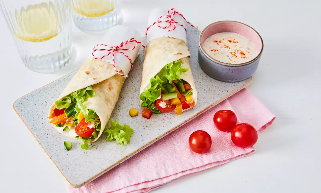 Gemüse-Wraps mit frischem Salat, roten Tomaten und bunten Paprikastückchen auf heller Platte mit rosa Stofftuch und Dip in Schale