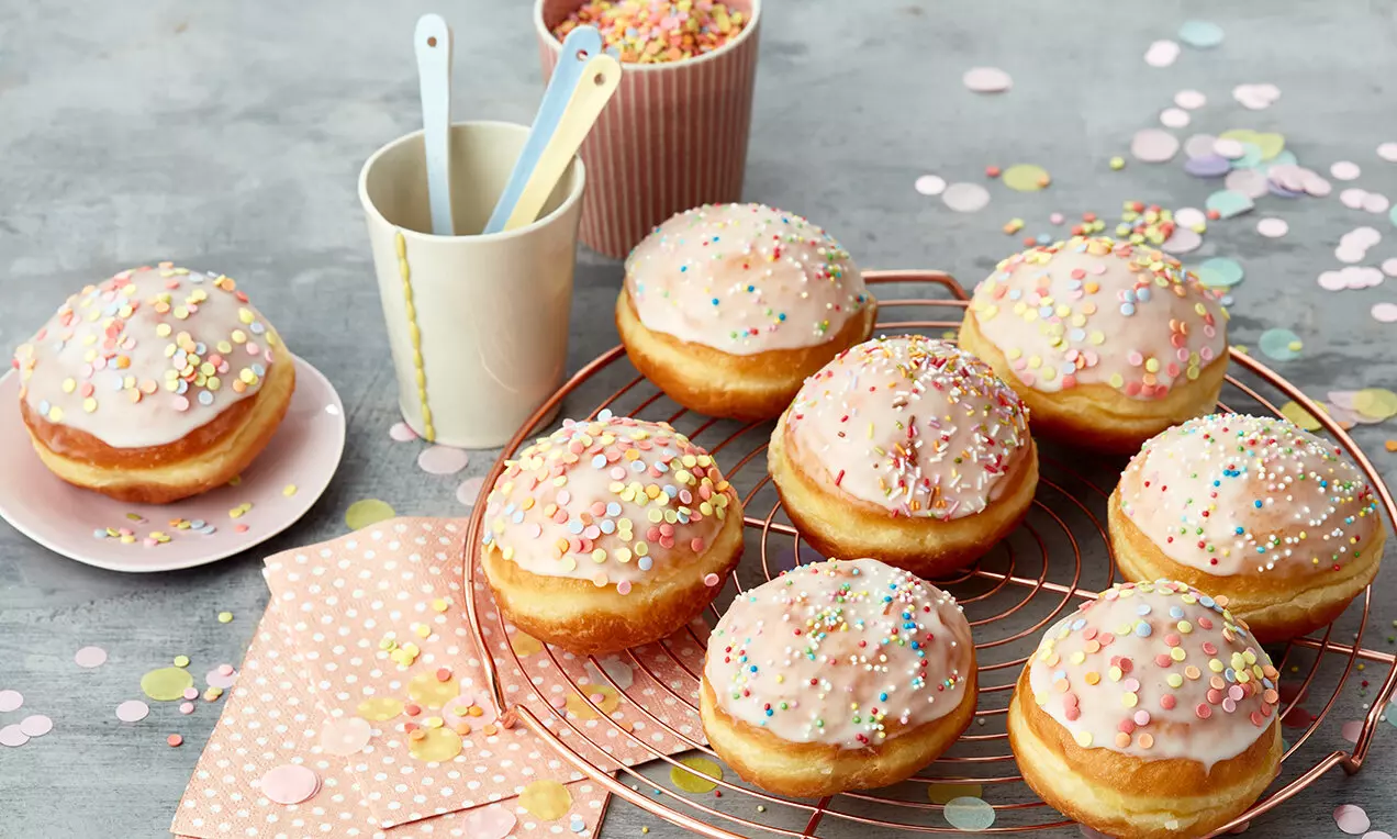 Berliner dorati e soffici con glassa rosa lucida, decorati con piccoli confetti colorati, disposti su una griglia rotonda sopra un tavolo grigio