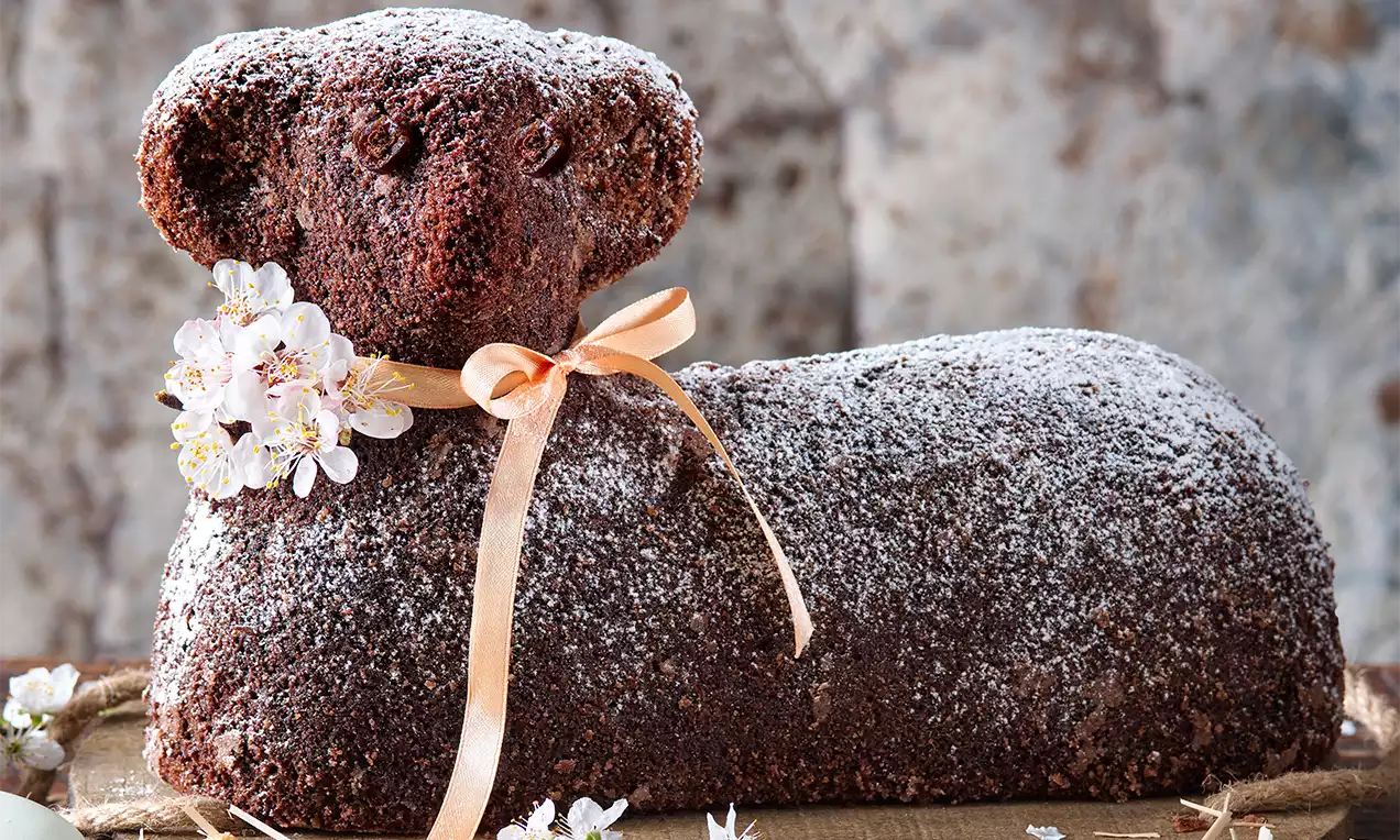 Kakaový baranček tmavohnedý s jemne zrnitou textúrou, posypaný práškovým cukrom, ozdobený mašľou a kvetmi na prírodnom podklade