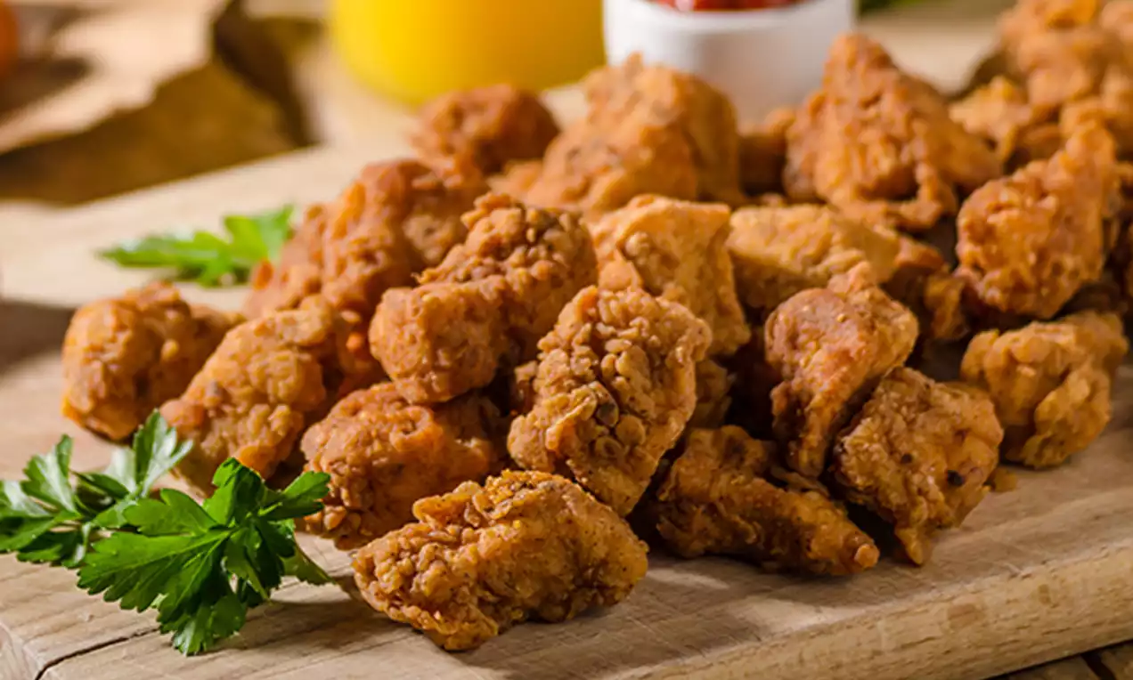 Golden brown crispy chicken bites with a rough texture arranged on a wooden board, garnished with parsley and served near dipping sauces