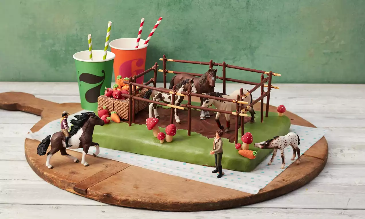 Rechteckiger Kuchen mit grüner Marzipandecke, dekoriert als Pferdekoppel mit braunen Zäunen, Figuren und bunten Zucker-Obststücken