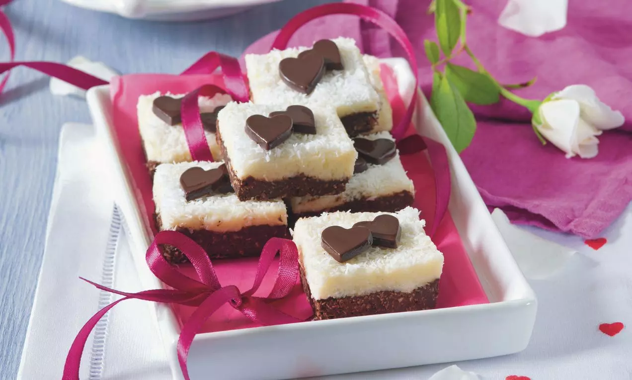 Kostky s bielou kokosovou vrstvou a tmavým čokoládovým základom, ozdobené čokoládovými srdiečkami na ružovom podklade v bielej miske