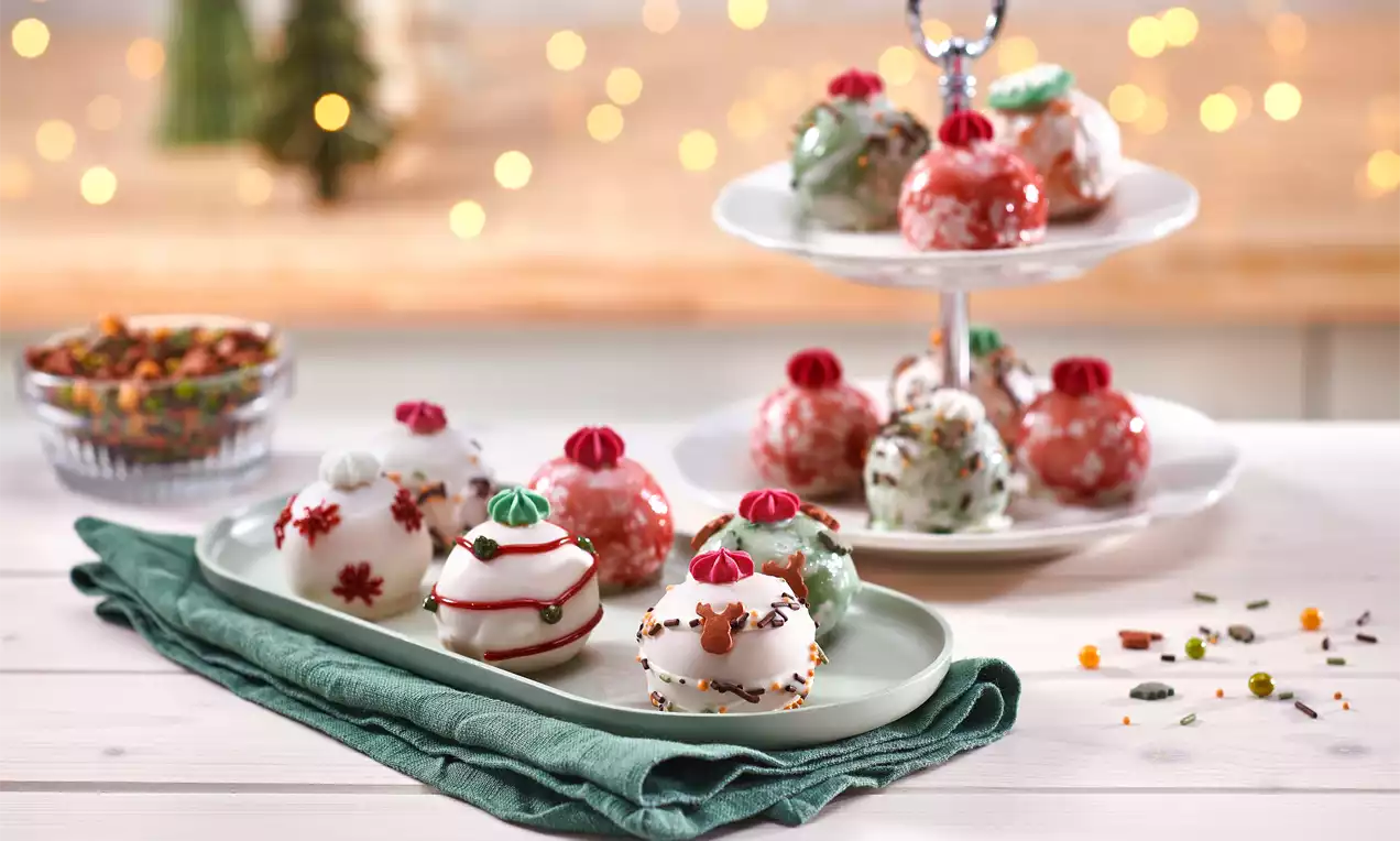 Festlich dekorierte, glänzende Cakepops in Rot, Weiß und Grün mit Zuckerguss und Streuseln auf Teller und Etagere vor unscharfer Lichterkulisse