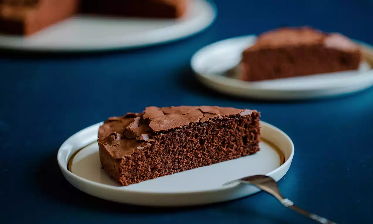 Dış yüzeyi hafif çatlak, nemli ve kabarık dokulu bir dilim çikolatalı kek, beyaz bir tabakta mavi arka plan üzerinde sunulmuş