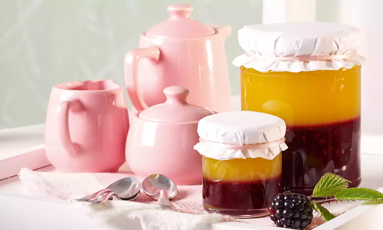 Zweischichtige Brombeer-Mango-Konfitüre in gläsernen Schraubgläsern mit weißem Deckel auf rosa Stoff, umgeben von pinkem Porzellan und Besteck