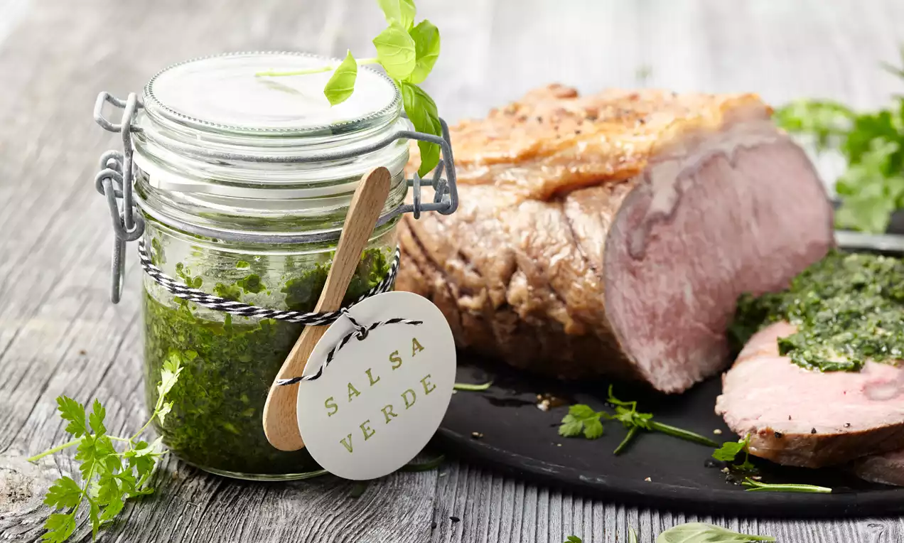 Grünes Salsa-Verde in einem Glas mit Holzlöffel, daneben saftiger Braten und Scheiben mit Kräutersoße auf schwarzem Teller mit frischen Kräutern