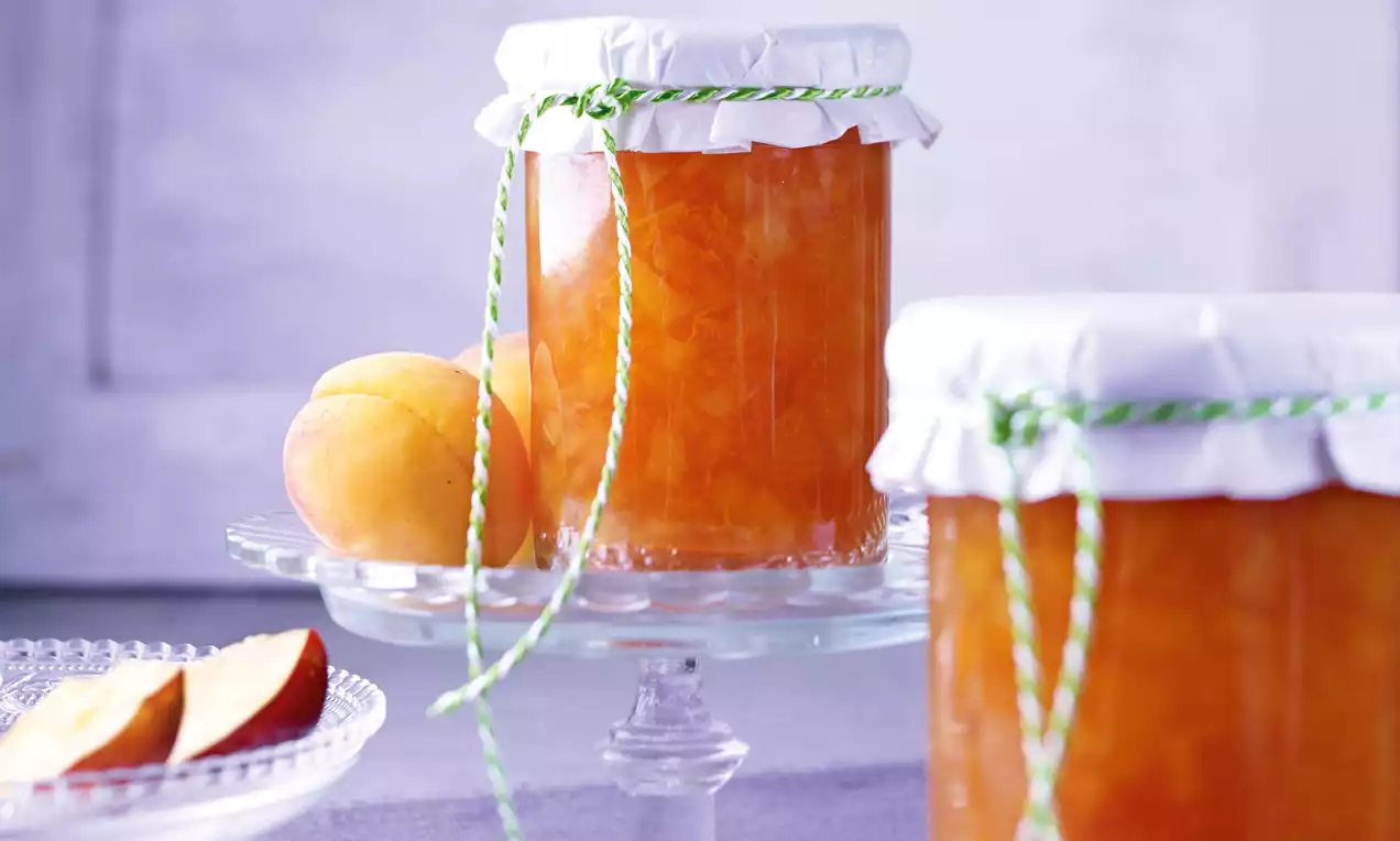 Pots de confiture ambrée aux reflets dorés, couvercles blancs bordés de ficelle verte, disposés sur un présentoir en verre avec des abricots et des tranches sur une assiette transparente