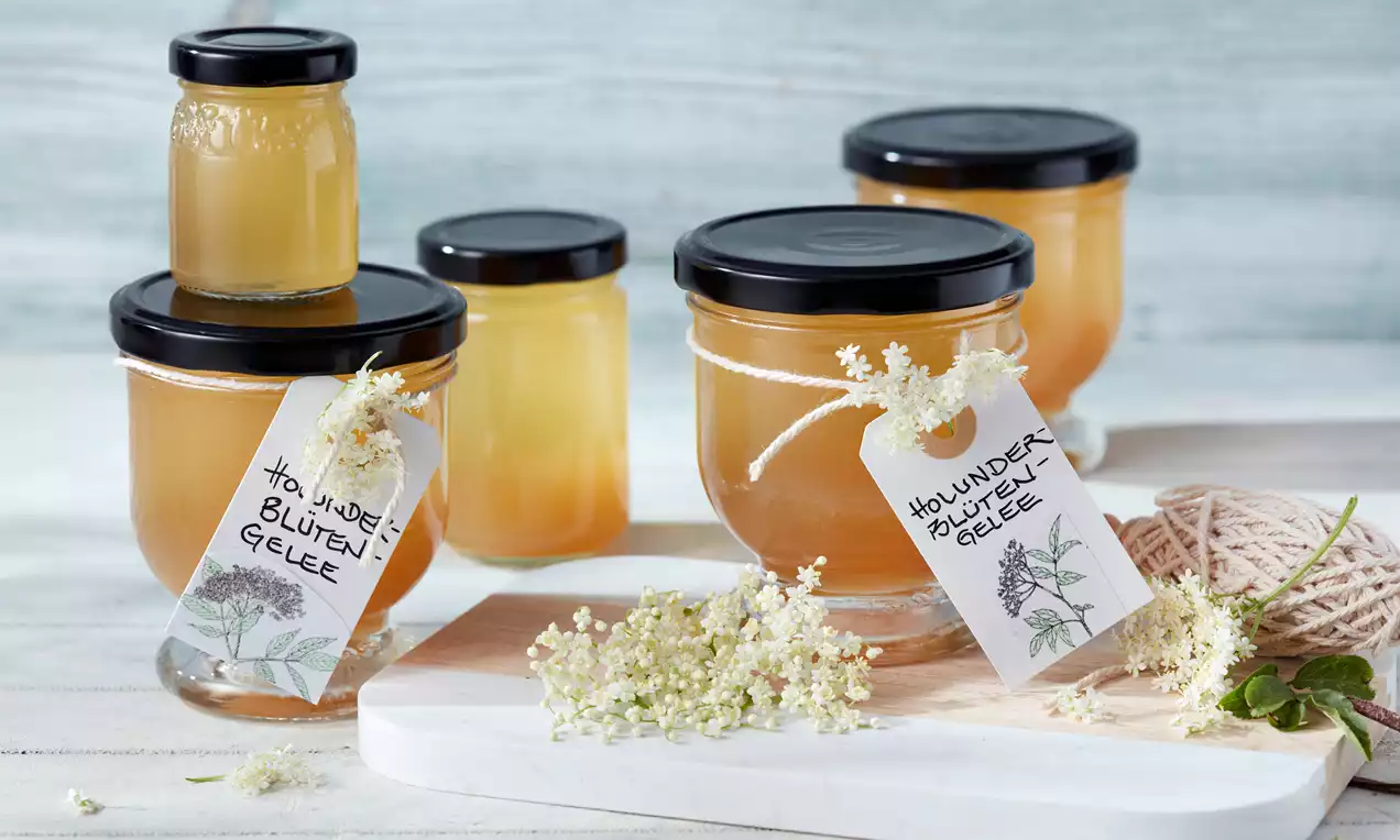 Goldgelbes Holunderblüten-Gelee in Gläsern mit schwarzen Deckeln und handbeschrifteten Anhängern, dekoriert mit frischen Holunderblüten