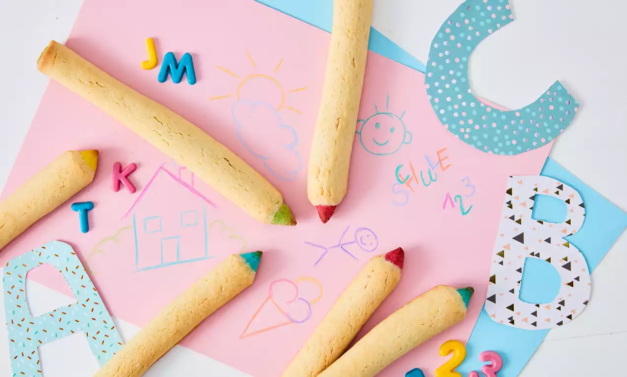 Essbare Buntstifte aus hellbraunem Gebäck mit farbigen Spitzen, arrangiert auf buntem Papier mit gemalten Zeichnungen und Deko-Buchstaben