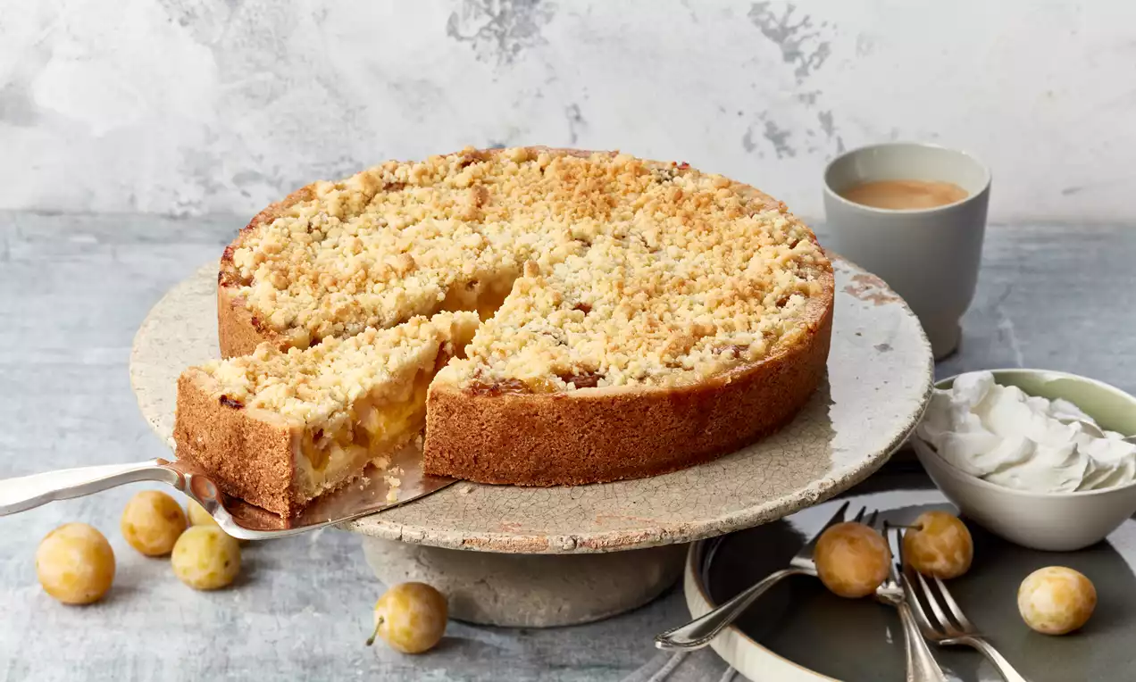 Goldgelber Mirabellenkuchen mit knusprigen Streuseln auf grauer Tortenplatte, begleitet von Mirabellen, Sahne und Teetasse in heller Umgebung