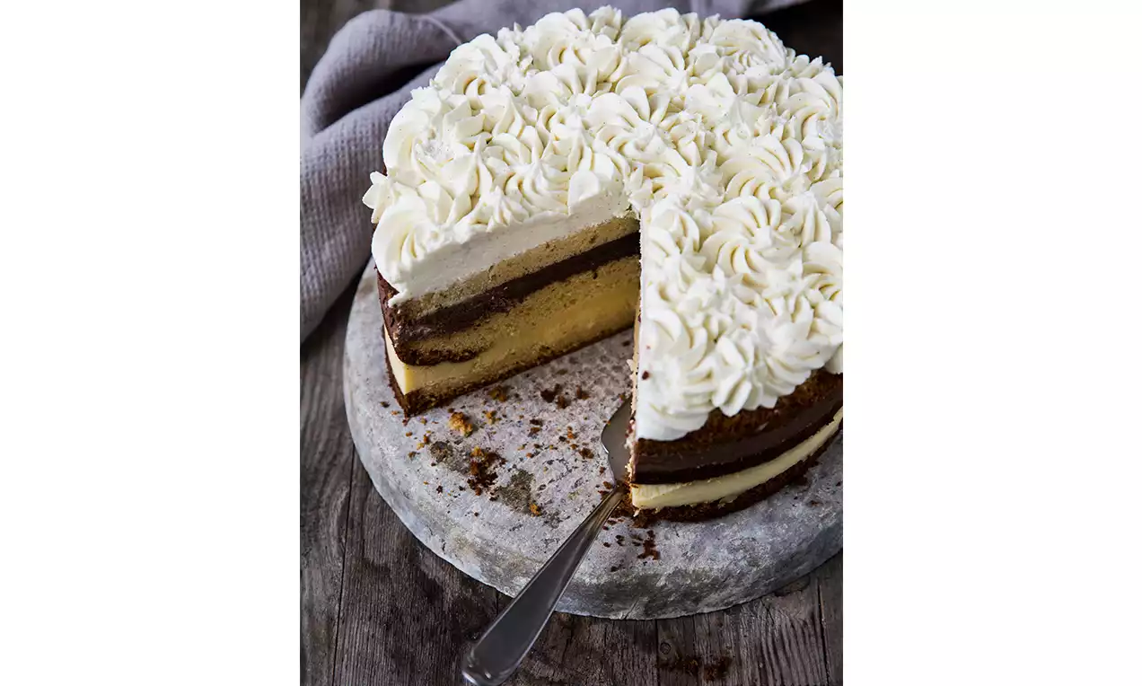 Torta stratificata con base scura, crema dorata e soffice, decorata con riccioli di panna bianca su un piatto rustico di marmo grigio