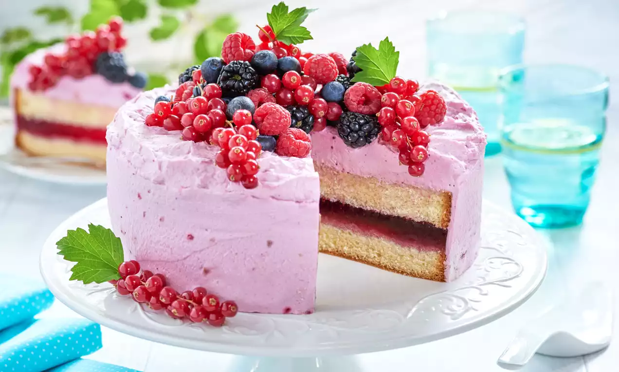 Tort z warstwą biszkopta i galaretki, pokryty różową masą śmietanową, udekorowany owocami leśnymi i zielonymi listkami na białej paterze