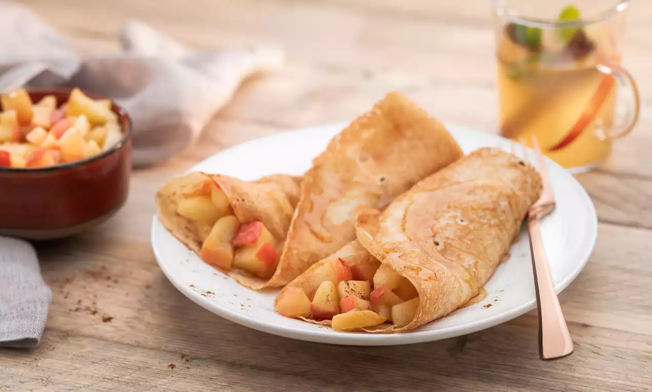 Drie goudbruine pannenkoeken gevuld met warme appelstukjes, geserveerd op een wit bord met een kop heldere thee en een kommen appelvulling