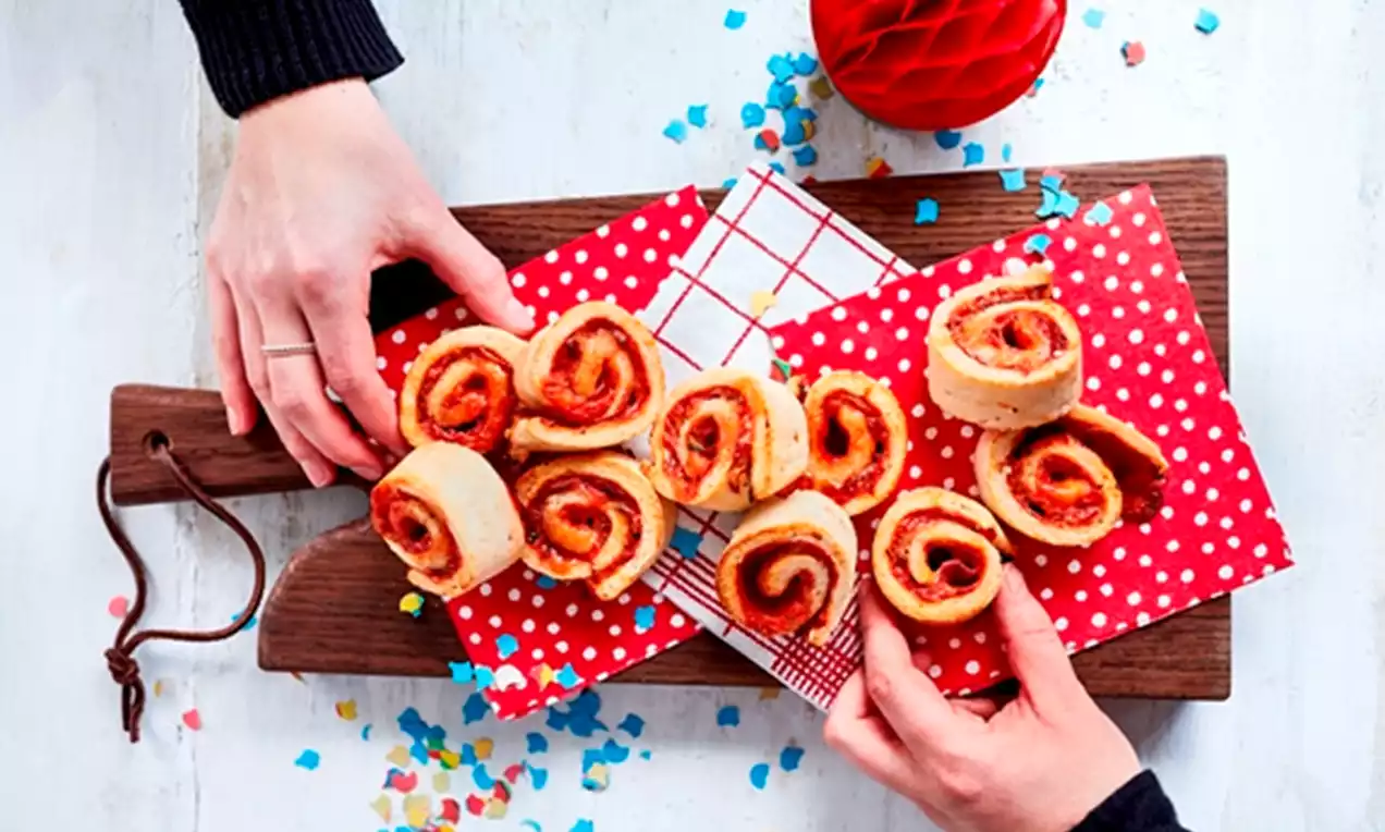 Golden brown pizza swirls with spiral layers of red tomato sauce and melted cheese on patterned red napkins atop a wooden serving board