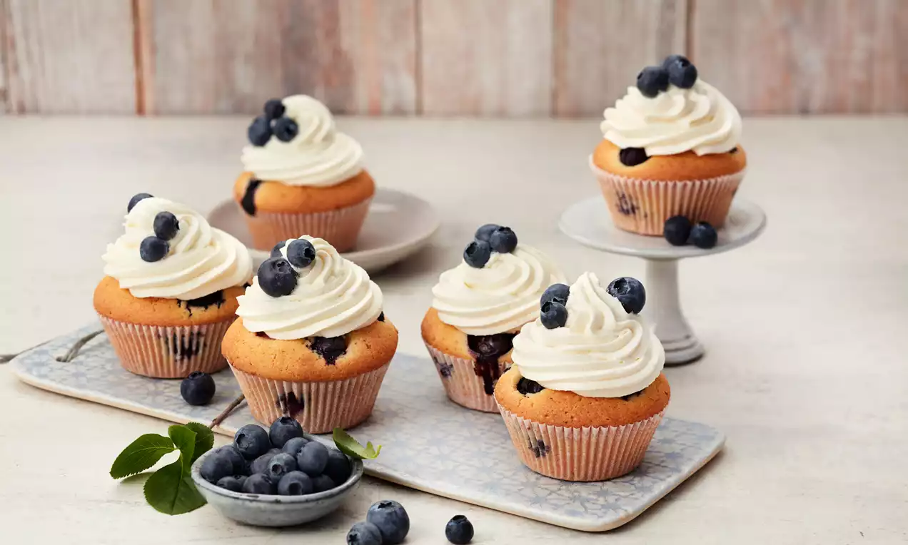 Goldene Blaubeer-Cupcakes mit weißer Sahnehaube, frischen Blaubeeren dekoriert, auf Tabletts und einer Tortenplatte arrangiert