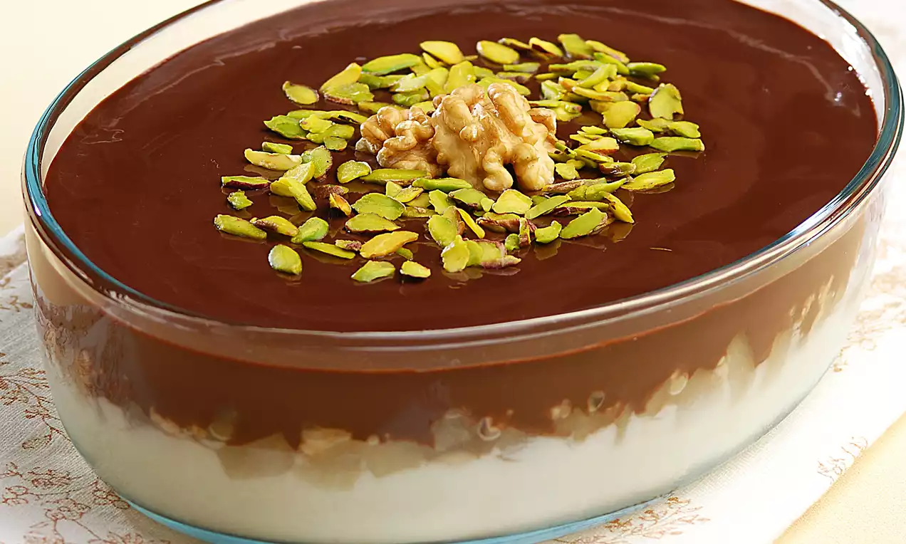 Oval cam kapta beyaz sütlü tatlı üzerine parlak çikolata sosu, ortasında ceviz ve ince kıyılmış yeşil fıstıklarla süslenmiş