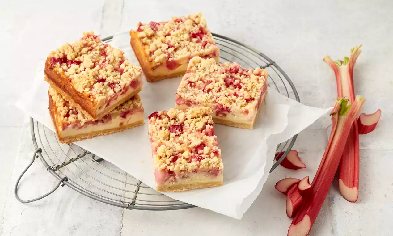 Rhabarber-Quarkkuchen in rechteckigen Stücken mit goldbrauner Streuselschicht auf weißem Backpapier, daneben frische Rhabarberstangen