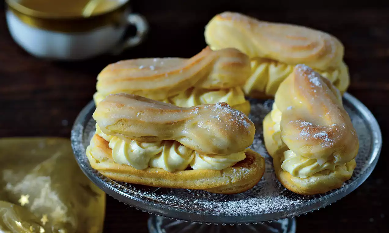 Podlouhlé nadýchané éclairs zlatavé barvy plněné světlým krémem, posypané moučkovým cukrem na skleněném podnosu