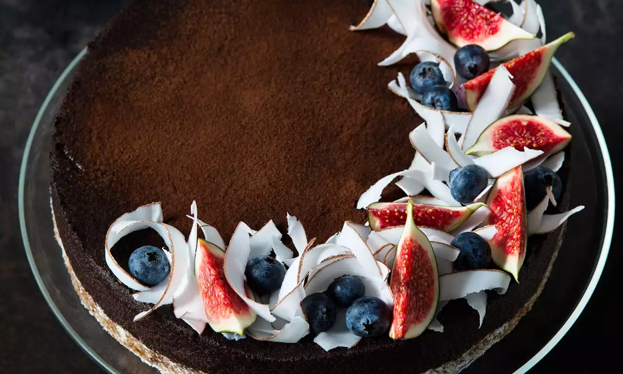 Čokoládově hnědý karobový cheesecake sypaný kakaovým práškem, ozdobený fíky, borůvkami a tenkými kokosovými plátky na skleněném talíři