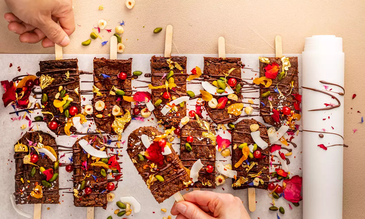 Donkerbruine suikervrije brownieplakken op een houten stok, bedekt met kleurrijke bloemen, noten, kokosschilfers en gedroogd fruit op bakpapier
