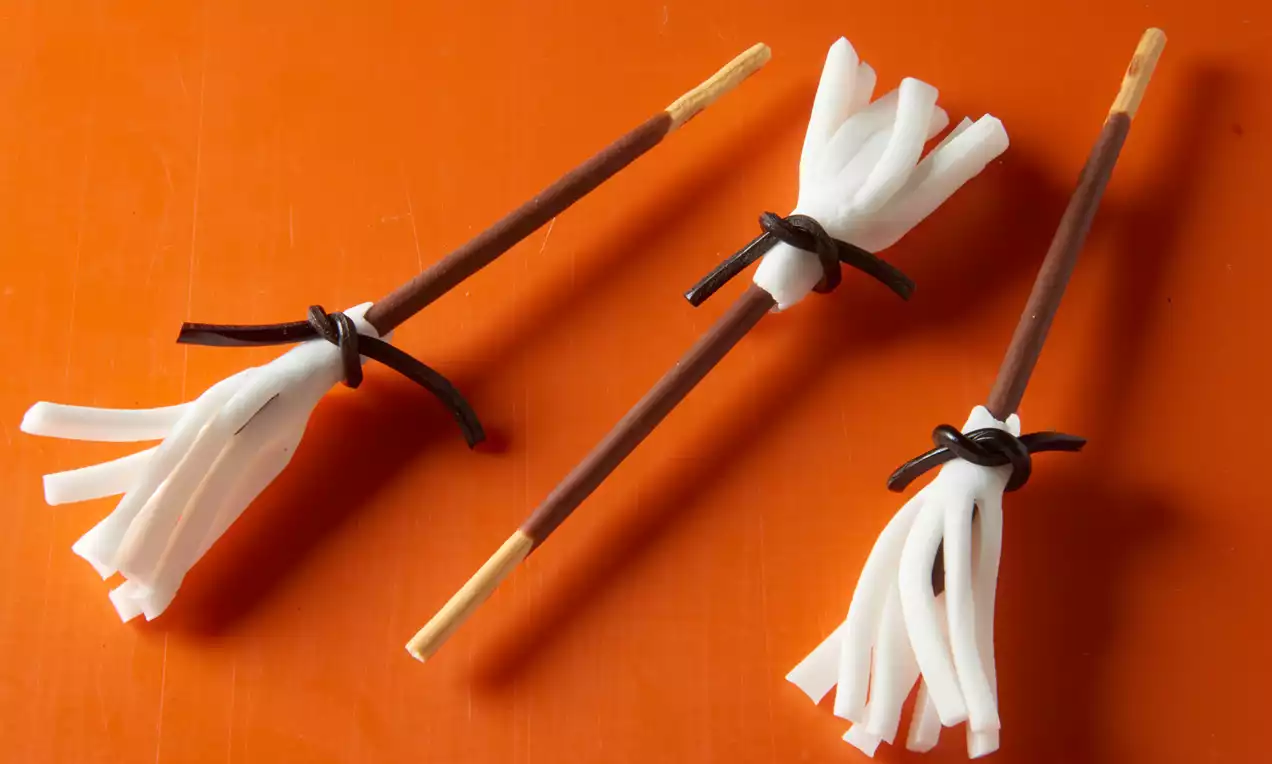 Drei kleine essbare Hexenbesen aus braunen Schokostäbchen mit weißen Fransen aus weicher Zuckermasse und schwarzen Schleifen auf orangefarbenem Untergrund