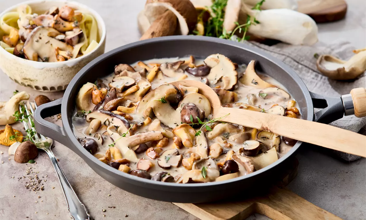 Pfanne mit Champignons, Waldpilzen, Zwiebeln in cremiger Sauce mit Pfannenwender, daneben Waldpilze, Besteck, Nudeln – schnelles, herzhaftes Rezept für vegetarisches Pfannengericht oder Beilage.
