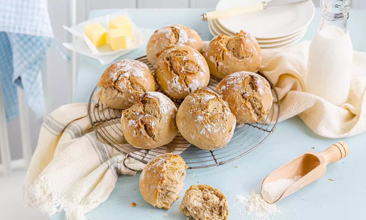 Goldbraune Sauerteig-Brötchen mit rissiger Kruste liegen auf einem runden Gitter, umgeben von Butterwürfeln, Mehl und Milchflasche
