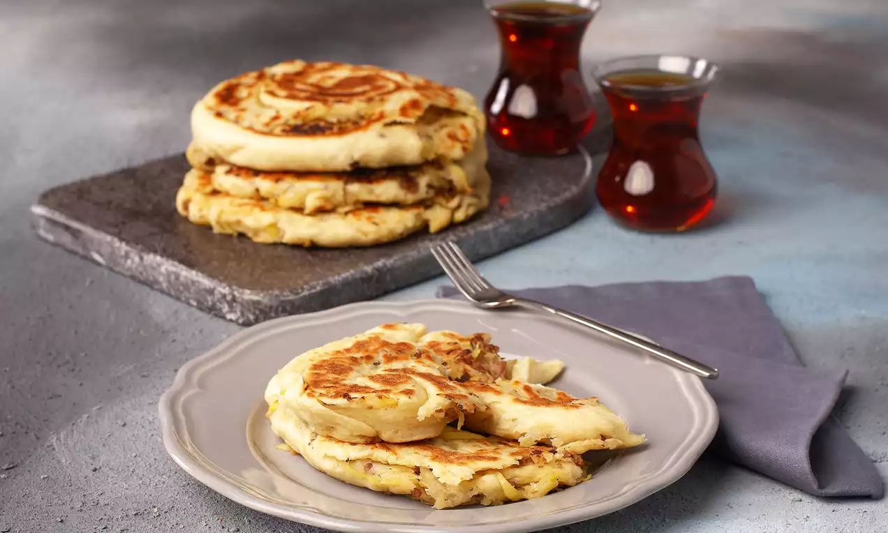 Altın rengi kızarmış patatesli kıymalı bazlama, sade gri tabakta dilimlenmiş, yanında iki bardak çay ve gri bir peçete ile sunulmuş