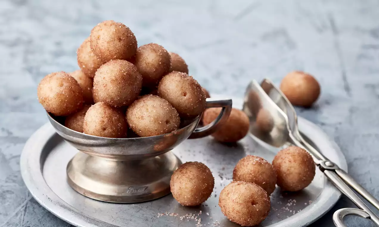 Kleine, rundliche Marzipankartoffeln mit goldbrauner Zuckerhülle in einer silbernen Schale auf einem Teller, daneben Zuckerzange