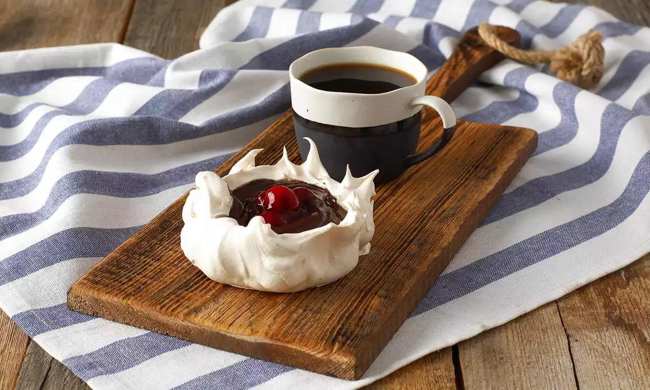 Kar beyazı kıtır pavlova tabanı, koyu çikolatalı supangle dolgusu ve tepesinde parlak kırmızı vişne ile ahşap sunum tahtasında.