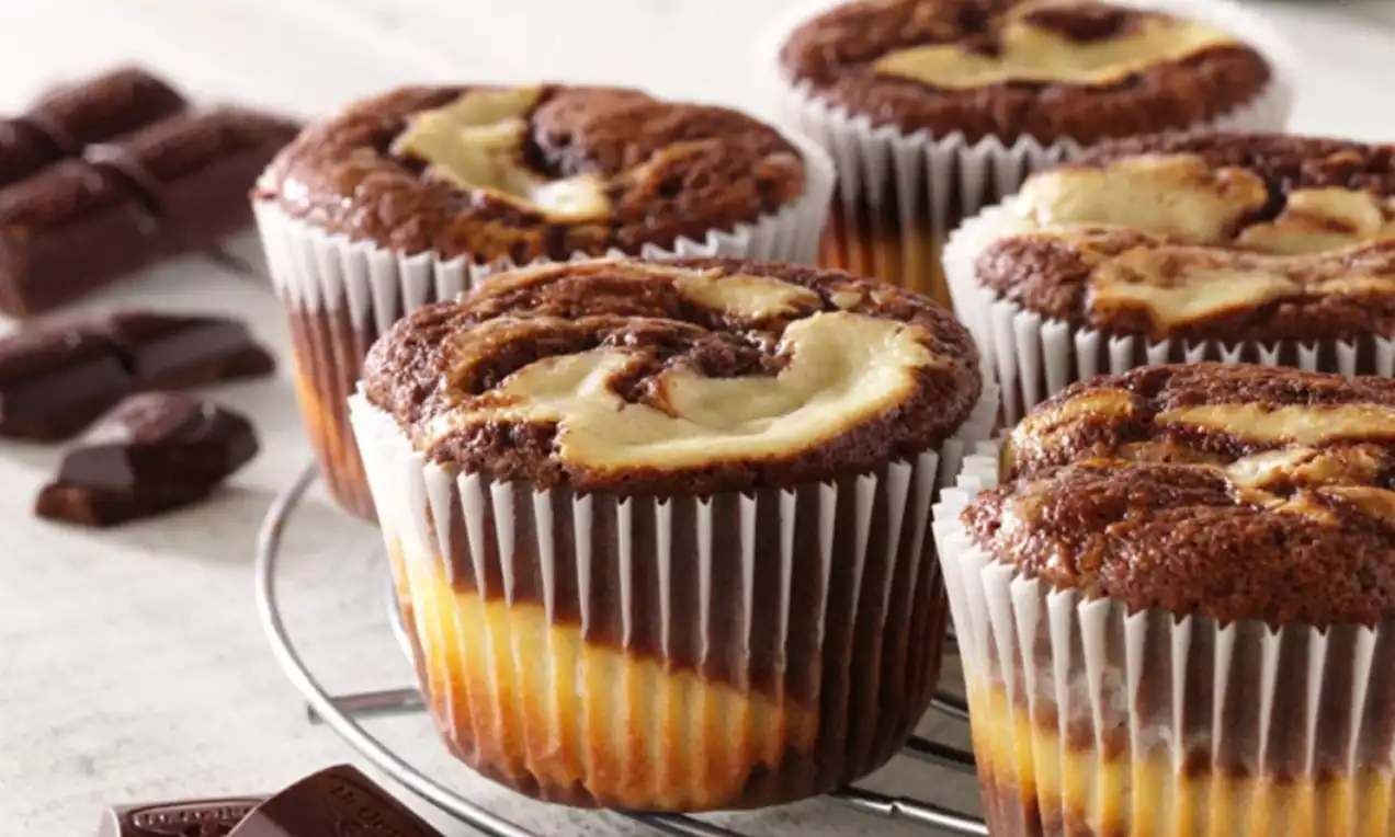 Marble chocolate cheesecake muffins with swirls of creamy beige and rich brown in ridged paper cases on a wire rack, surrounded by chocolate pieces