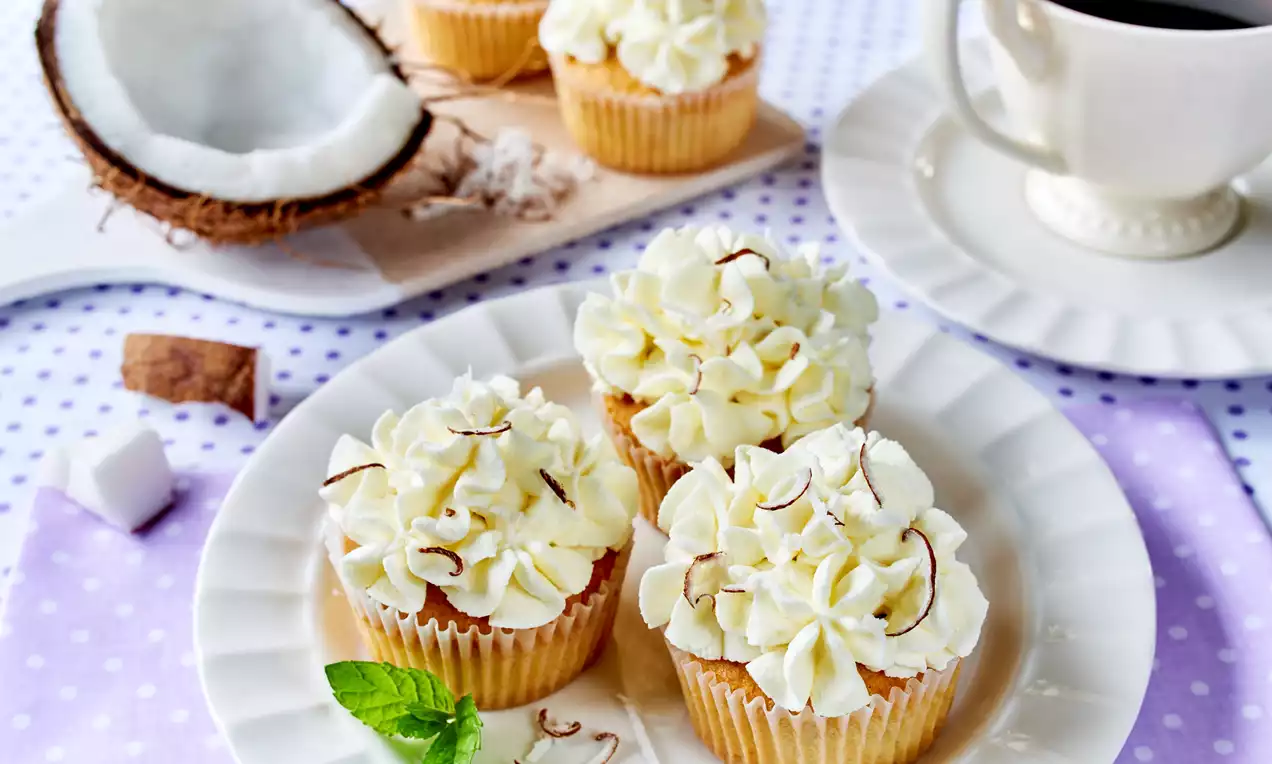 Kokosowe babeczki z kremową masą śmietanową, ozdobione wiórkami kokosowymi, na białym talerzu z listkiem mięty na fioletowym tle w grochy